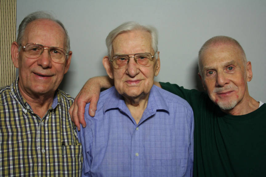 Three older white men stand arm in arm against a light colored backdrop