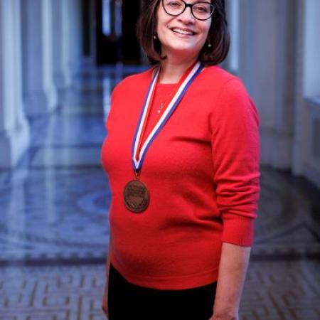 A vertical shot of author Meg Medina, standing and wearing the National  Ambassador for Young People's Literature Medal. She wears glasses and has shoulder length straight brown hair. She is wearing a red sweater and black pants. 